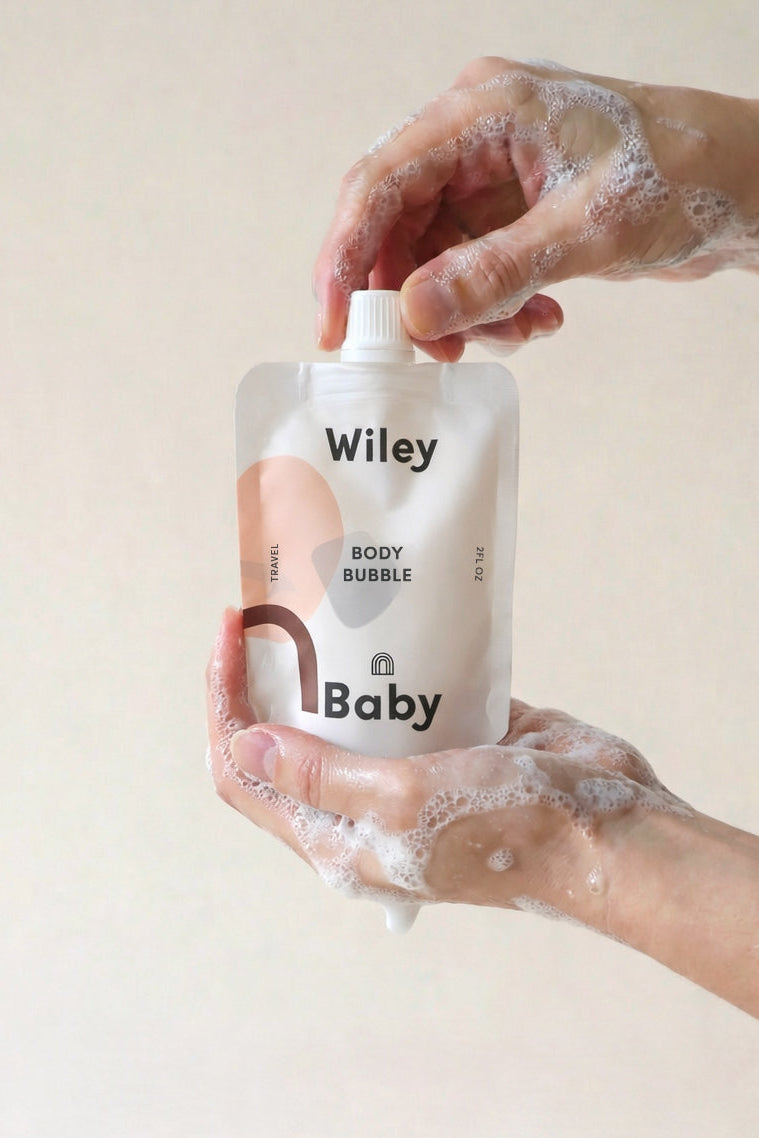 Hand holding a Wiley Baby body bubble product with soapy hands on a beige background