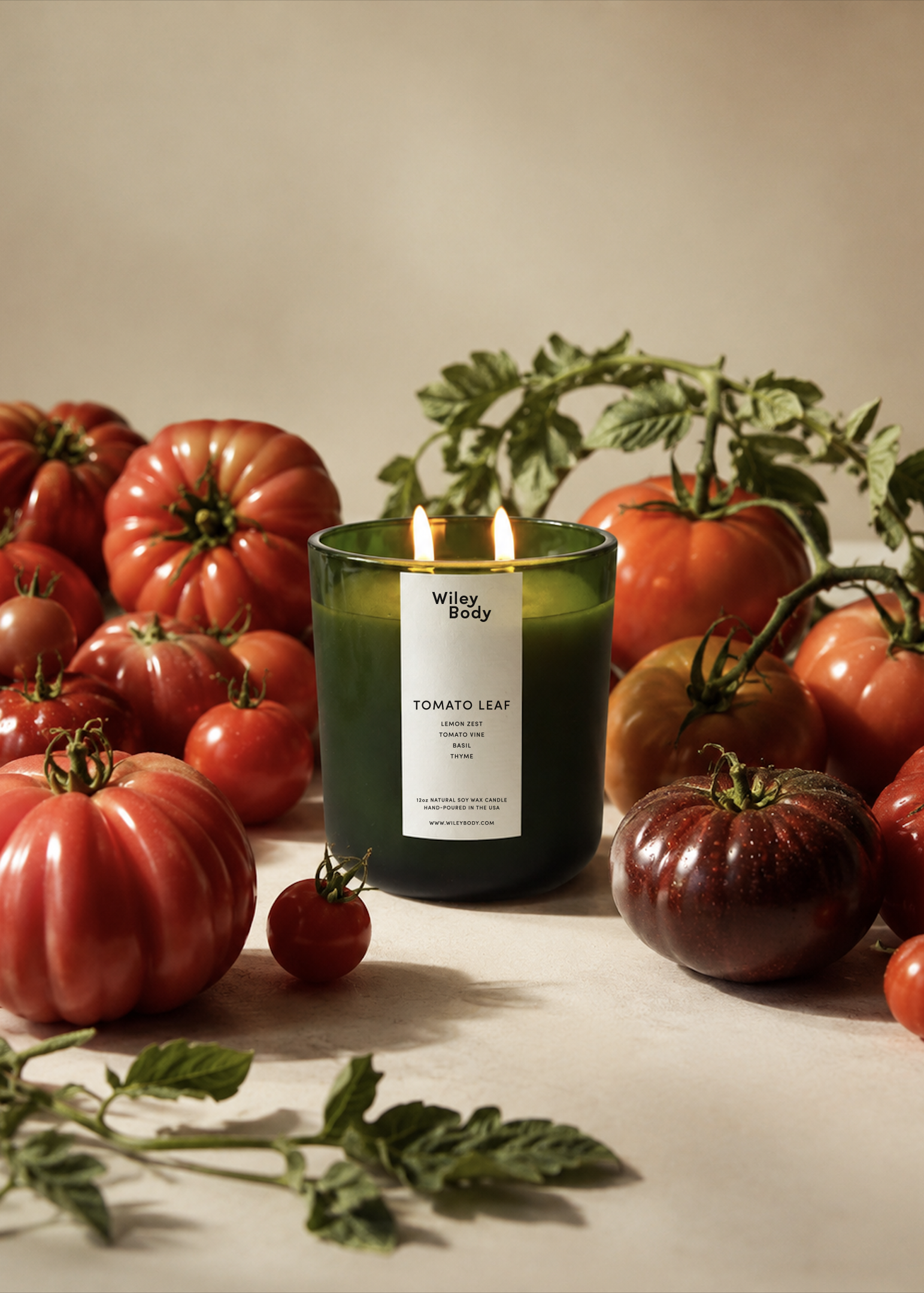 Wiley Body Tomato Leaf candle in a dark green glass vessel with a double wick, surrounded by heirloom tomatoes and fresh tomato vines on a neutral surface.